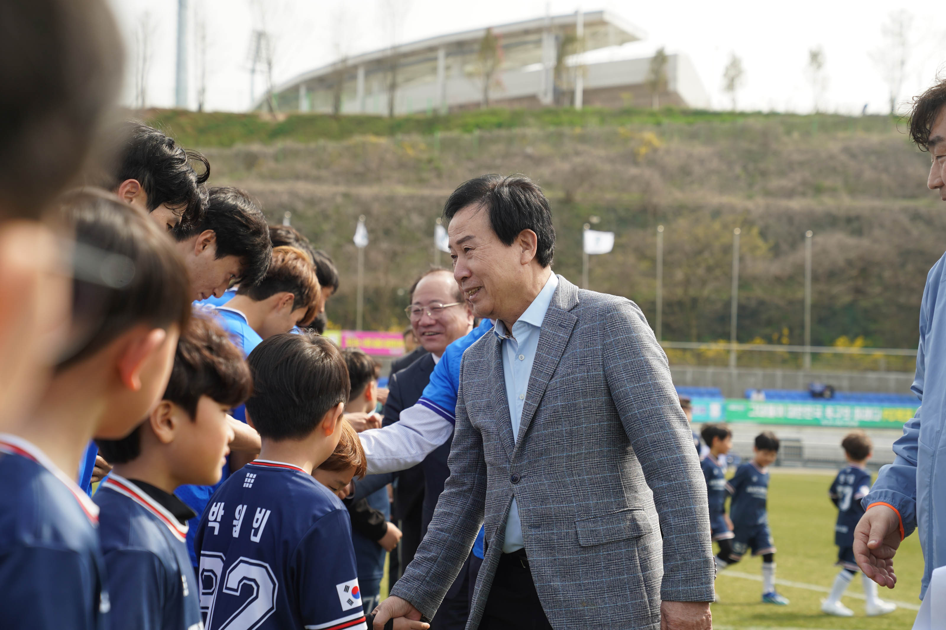 일렬로 줄지어 있는 파란색 유니폼을 입은 선수들 앞에 남색 유니폼을 입은 어린이들이 서있다. 그들과 악수를 하며 인사를 나누는 박홍률 시장의 사진