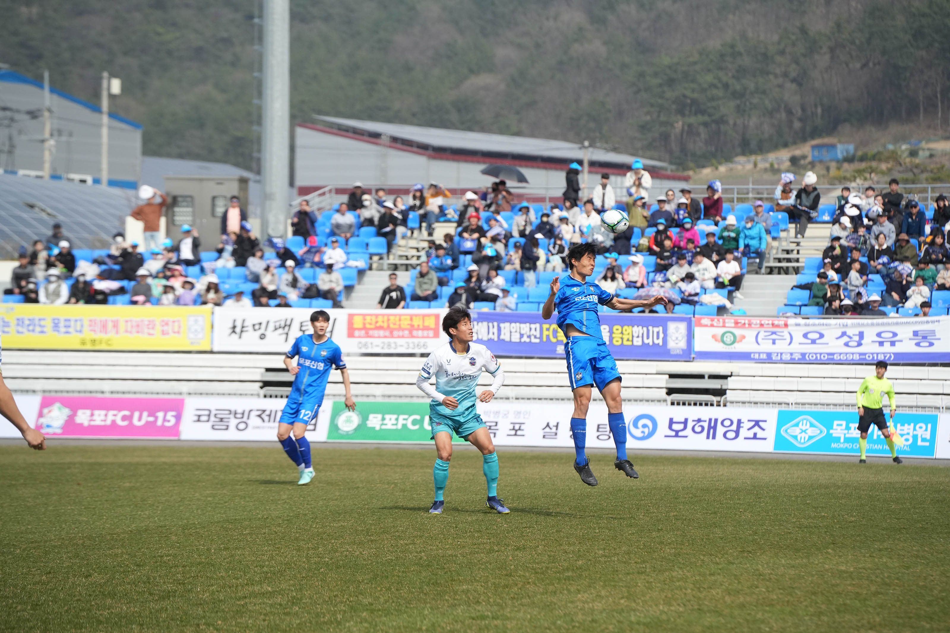 축구경기 모습 중 찍힌 사진. 세명의 선수들과 한명의 심판이 있고, 파란색 유니폼을 입은 선수가 헤딩하는 순간을 포착한 사진.