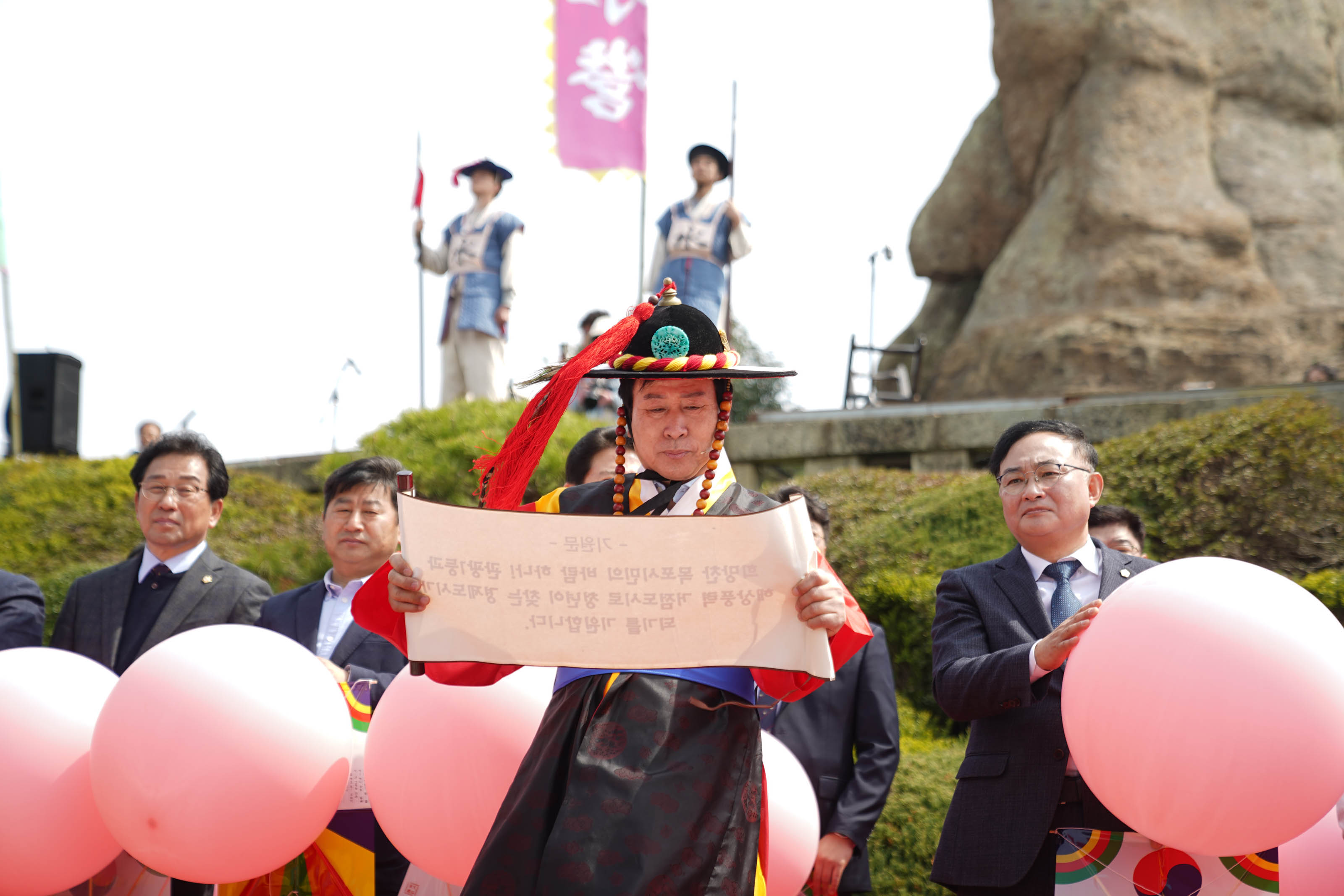 사또 분장을 한 박홍률 시장이 기원문 족자를 들고 읽고 있으며 그 뒤로 중년 남성의 참석내빈들이 연분홍색 커다란 풍선을 들고 서 있다.