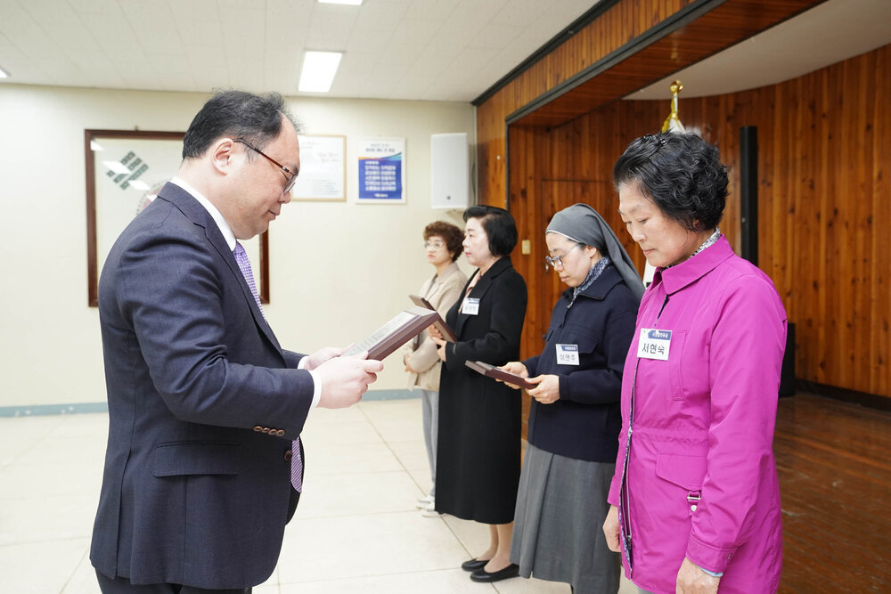 4월 정례조회에서 정장을 입은 남성이 분홍색 외투를 입은 여성에게 표창장을 전달하는 모습