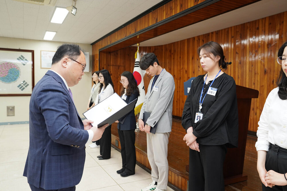 정장을 입고 안경을 낀 남성이 검은색 티를 입은 여성에게 상장을 수여하고 있다