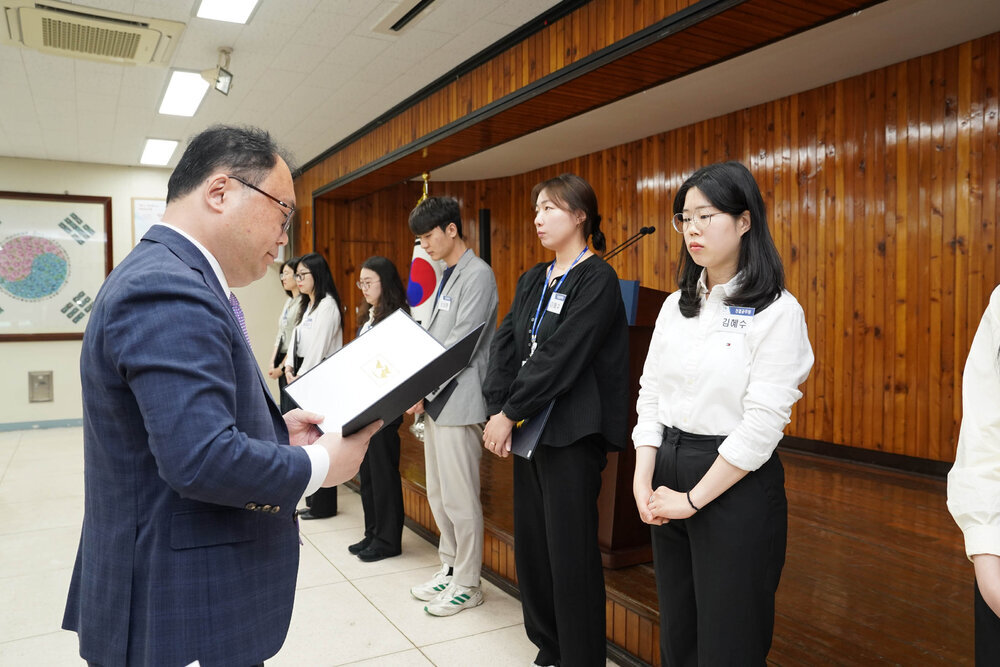 정장을 입고 안경을 낀 남성이 흰색 셔츠를 입은 여성에게 상장을 수여하고 있다
