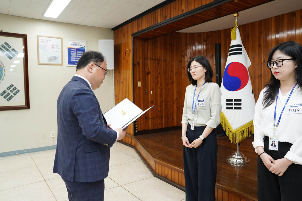정장을 입고 안경을 낀 남성이 연두색 셔츠를 입은 여성에게 상장을 수여하고 있다