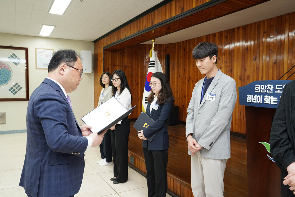 정장을 입고 안경을 낀 남성이 연한 회색 자켓을 입은 남성에게 상장을 수여하고 있다