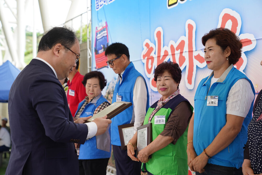 정장을 입고 안경을 낀 남성이 흰색 반팔 니트에 하늘색 작업 조끼를 입은 여성에게 패를 수여하고 있다