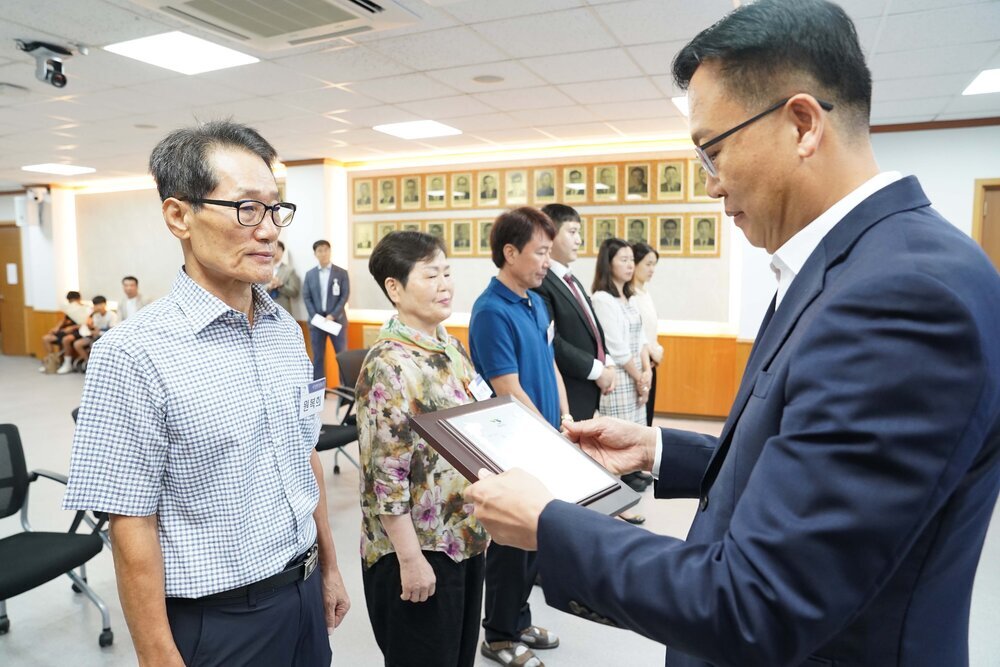 정장 차림의 관계자가 체크 셔츠를 입은 남성 수상자에게 표창패를 전달하며 내용을 읽고 있는 모습이다.