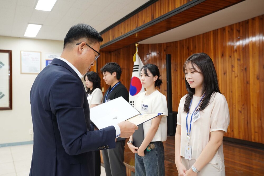 정장 차림의 관계자가 연베이지색 원피스를 입은 여성 수상자에게 패를 전달하며 읽고 있는 모습이다.