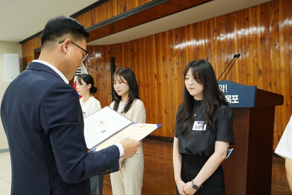 정장 차림의 관계자가 검은색 반팔 블라우스를 입은 여성 수상자에게 패를 전달하며 읽고 있는 모습이다.