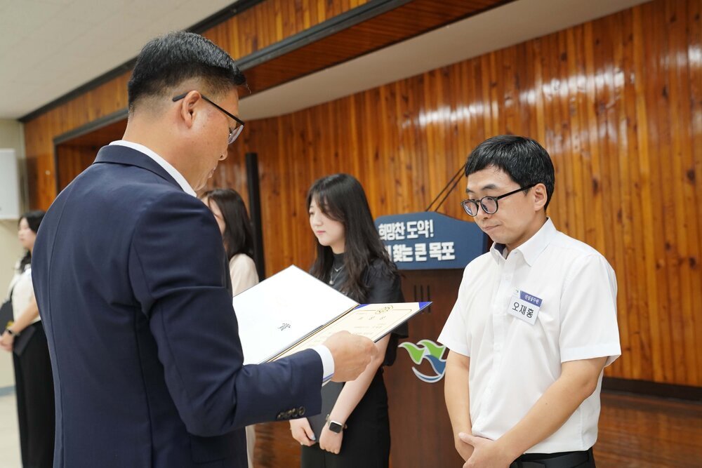 정장 차림의 관계자가 흰색 반팔 셔츠를 입은 남성 수상자에게 패를 전달하며 읽고 있는 모습이다.