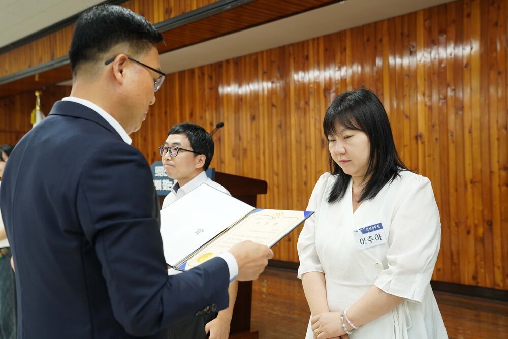 정장 차림의 관계자가 흰색 원피스를 입은 여성 수상자에게 패를 전달하며 읽고 있는 모습이다.