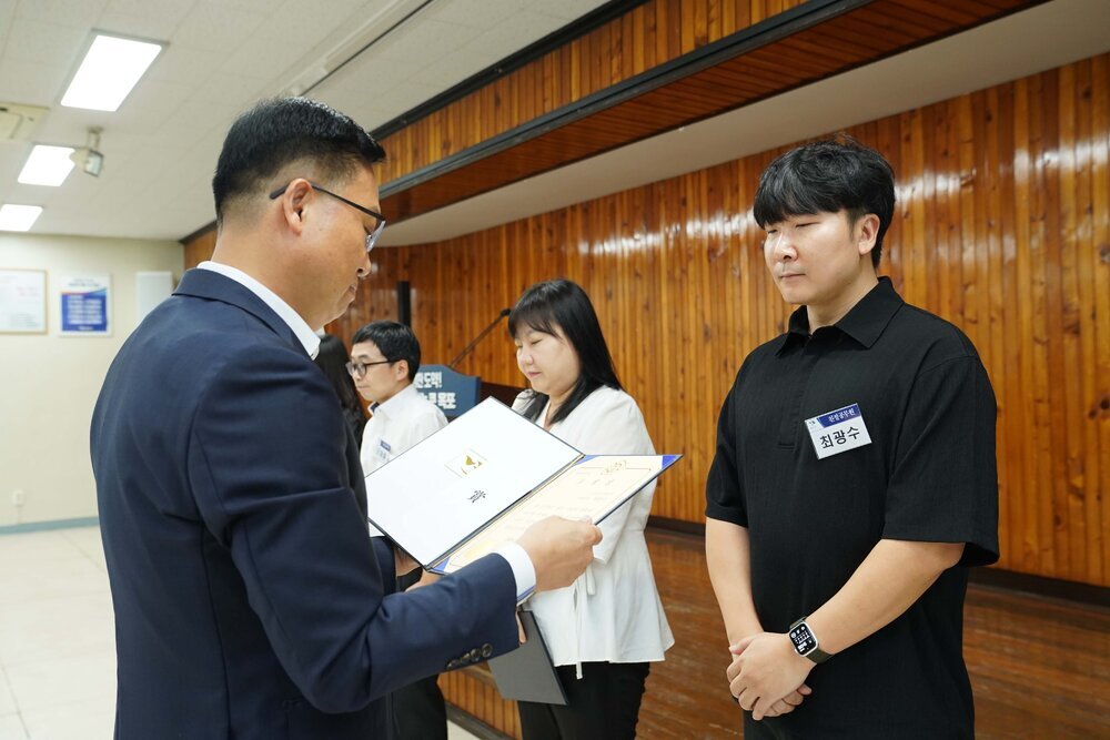 정장 차림의 관계자가 검은색 반팔 티셔츠를 입은 남성 수상자에게 패를 전달하며 읽고 있는 모습이다.