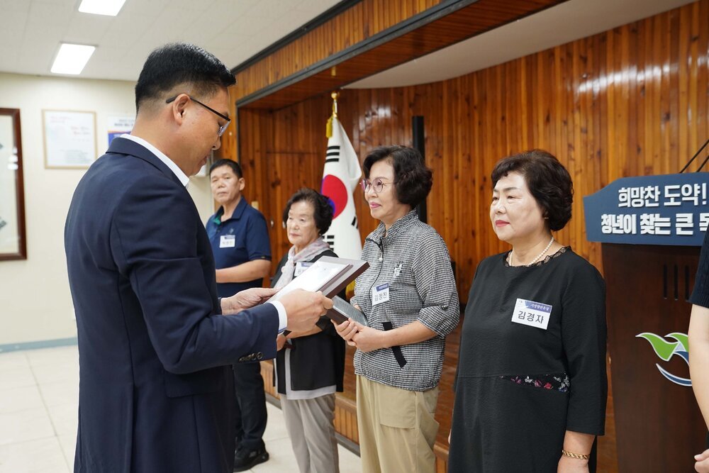 정장 차림의 관계자가 검은색 원피스를 입은 여성 수상자에게 패를 전달하며 읽고 있는 모습이다.