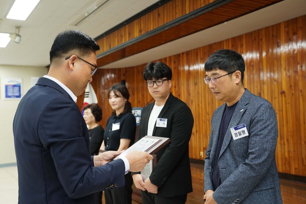 정장 차림의 관계자가 회색 재킷을 입은 남성 수상자에게 패를 전달하며 읽고 있는 모습이다.