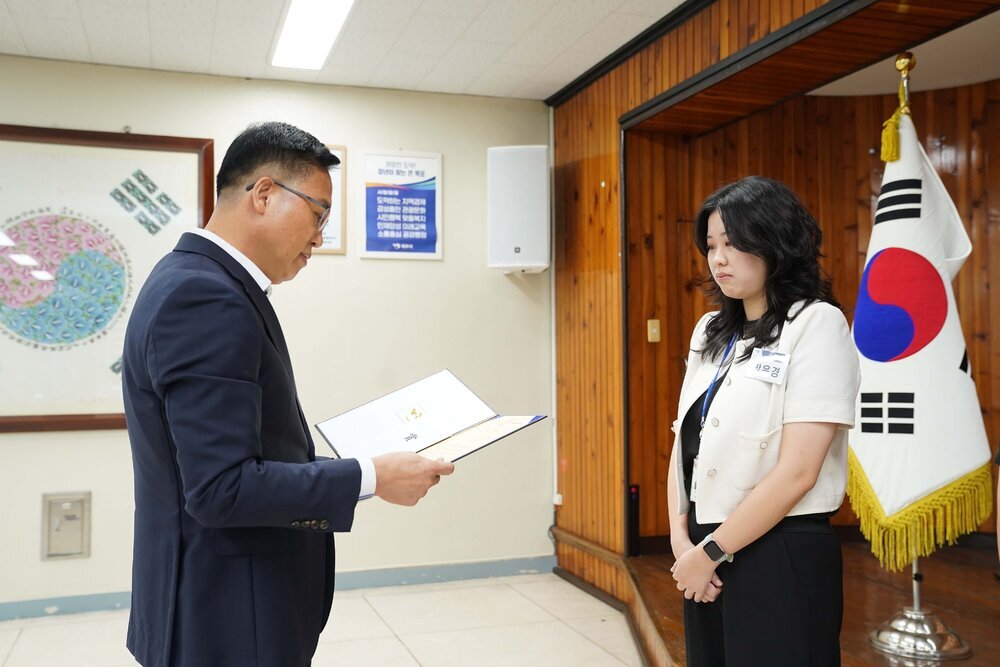 정장 차림의 관계자가 아이보리색 상의를 입은 여성 수상자에게 패를 전달하며 읽고 있는 모습이다.