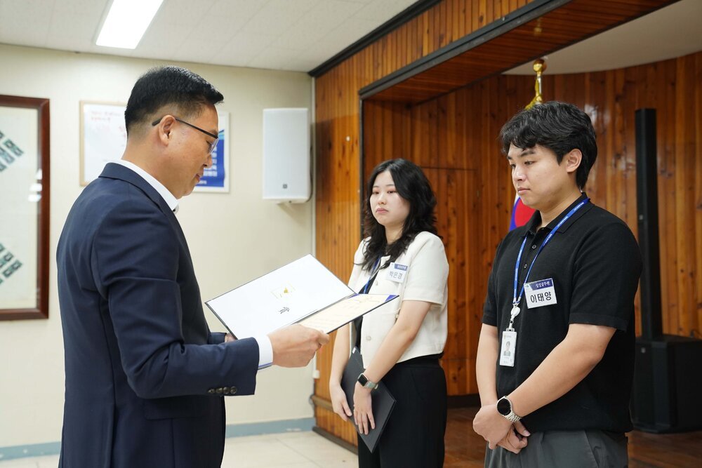 정장 차림의 관계자가 검은색 반팔 상의를 입은 남성 수상자에게 패를 전달하며 읽고 있는 모습이다.