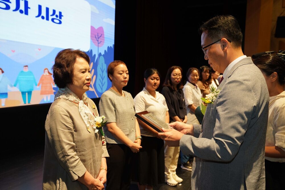 정장 차림의 관계자가 베이지색 재킷을 입은 여성 수상자에게 패를 전달하며 읽고 있는 모습이다.