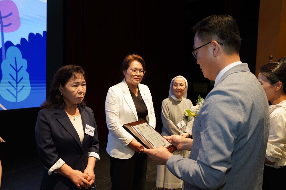 정장 차림의 관계자가 남색 재킷을 입은 여성 수상자에게 패를 전달하며 읽고 있는 모습이다.