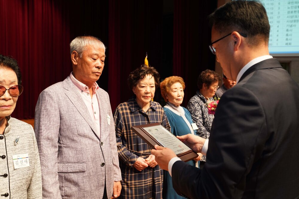 정장 차림의 관계자가 회색 재킷을 입은 남성 수상자에게 패를 전달하며 읽고 있는 모습이다.