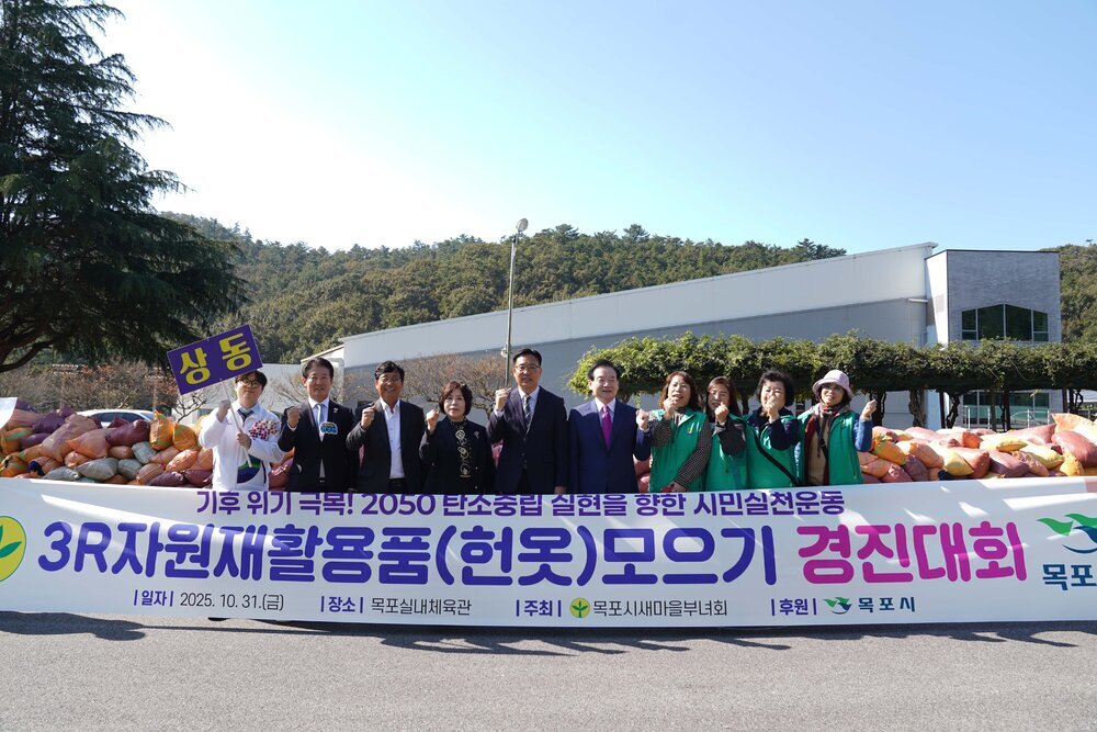 '3R 자원재활용품(헌옷) 모으기 경진대회' 현수막 뒤에서 상동 피켓을 든 참가자들이 파이팅 포즈를 취하고 있다. 쌓여있는 엄청난 양의 자원재활용품 자루들이 눈에 띈다.