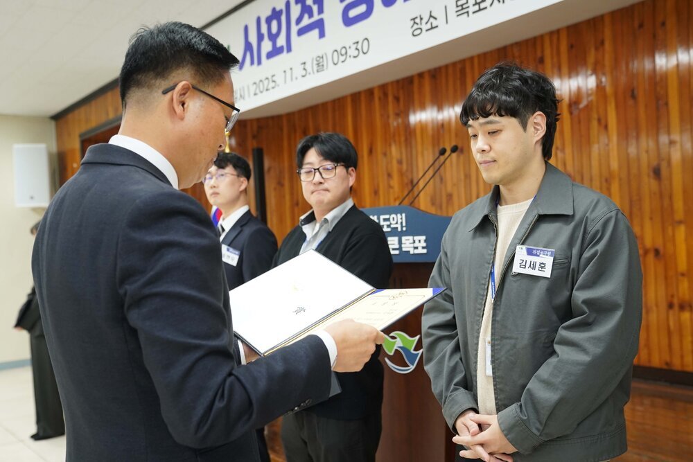 11월 정례조회로 정장을 입고 안경을 쓴 남성이 짧은 파마머리에 파스텔톤 카키색 상의에 김세훈 수상자에게 상장을 수여 하고 있다.
