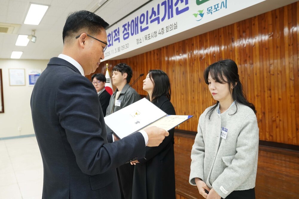 11월 정례조회로 정장을 입고 안경을 쓴 남성이 긴 곱슬머리를 반 뒤로 묵고서 밝은색 짧은 코트를 입은 조정선 수상자에게 상장을 수여 하고 있다.