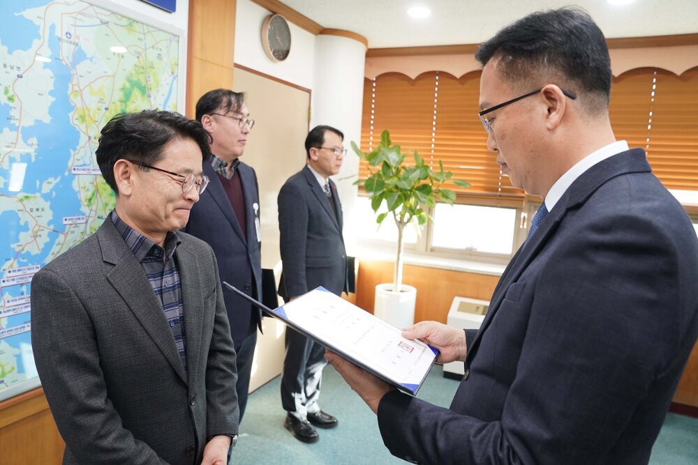 2026년 상반기 4급이하 정기인사로 정장을 입은 남성이 실내 사무실에서 한 남성에게 상장을 전달하며 내용을 읽고 있고, 남성은 안경을 쓰고 회색 체크무늬 자켓을 입은 채 두 손을 모으고 서 있으며 뒤쪽으로 다른 참석자들이 지켜보고 있다.