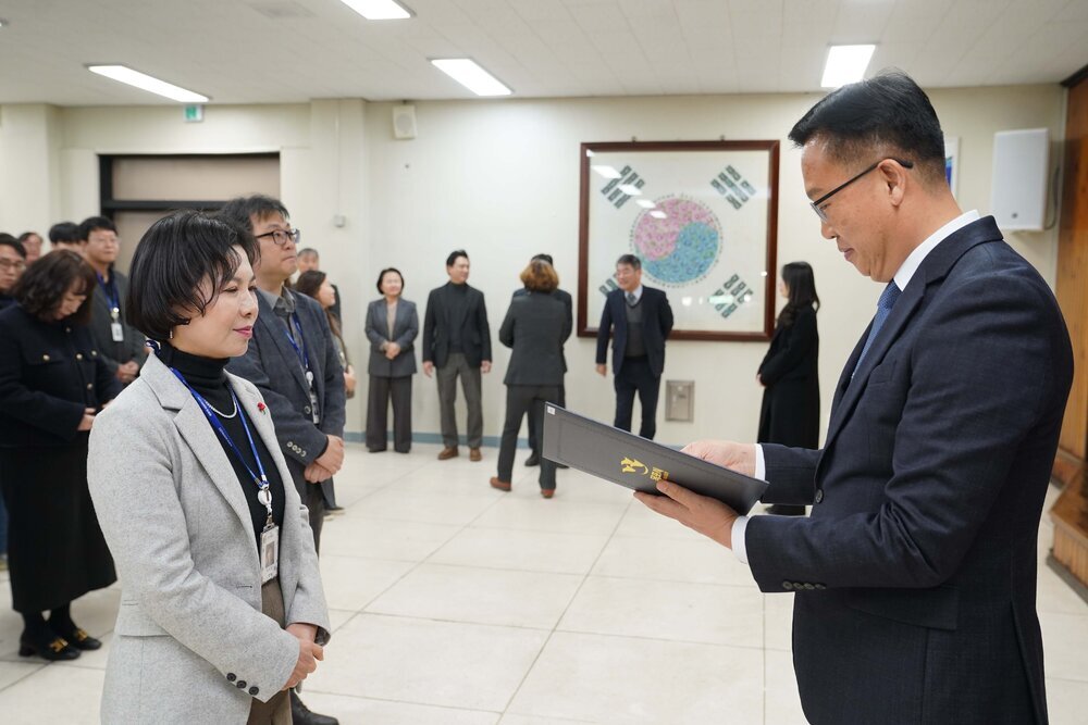 2026년 상반기 4급이하 정기인사로 정장을 입은 남성이 실내 행사장에서 한 여성에게 상장을 전달하며 내용을 읽고 있고, 여성은 검은색 목폴라 위에 밝은 회색 자켓을 입고 사원증을 착용한 채 두 손을 모은 상태로 서 있으며 뒤쪽으로 참석자들이 행사를 지켜보고 있다.