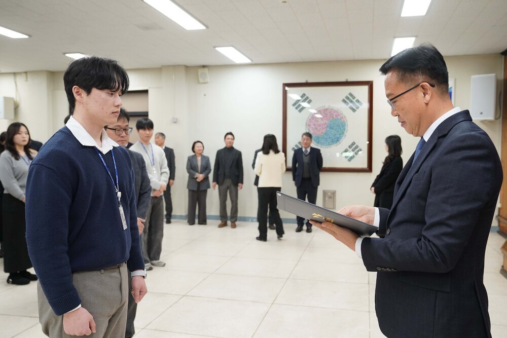 2026년 상반기 4급이하 정기인사로 정장을 입은 남성이 실내 행사장에서 한 남성에게 상장을 전달하며 내용을 읽고 있고, 남성은 흰 셔츠에 남색 니트를 겹쳐 입고 사원증을 착용한 채 두 손을 내린 상태로 서 있으며 뒤쪽으로 참석자들이 줄지어 서서 행사를 지켜보고 있다.