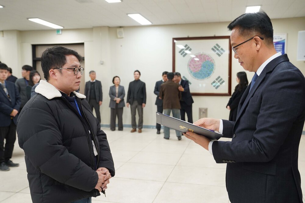 2026년 상반기 4급이하 정기인사로 정장을 입은 남성이 실내 행사장에서 한 남성에게 상장을 전달하며 내용을 읽고 있고, 남성은 안경을 쓰고 목 부분에 흰색 털이 있는 검은색 패딩을 입고 두 손을 모은 상태로 서 있으며 뒤쪽으로 참석자들이 줄지어 서서 행사를 지켜보고 있다.