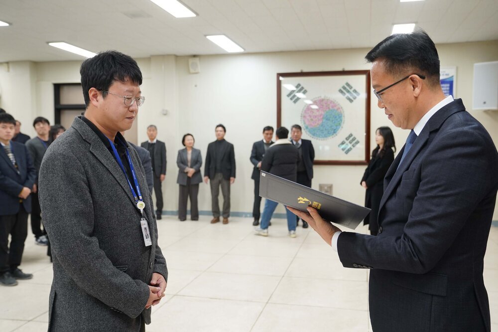 2026년 상반기 4급이하 정기인사로 정장을 입은 남성이 실내 행사장에서 한 남성에게 상장을 전달하며 내용을 읽고 있고, 남성은 안경을 쓰고 짙은 회색 보풀 니트 자켓을 입고 사원증을 착용한 채 두 손을 모은 상태로 서 있으며 뒤쪽으로 참석자들이 줄지어 서서 행사를 지켜보고 있다.