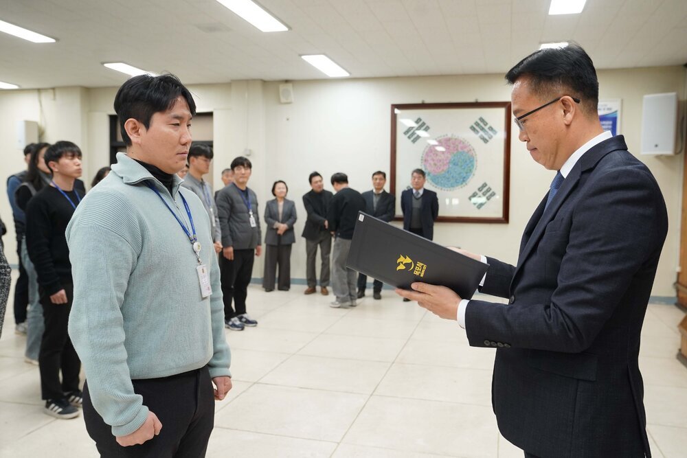 2026년 상반기 4급이하 정기인사로 정장을 입은 남성이 실내 행사장에서 한 남성에게 상장을 전달하며 내용을 읽고 있고, 남성은 연두색 니트를 입은 채 두 손을 모은 상태로 서 있으며 뒤쪽에는 참석자들이 줄지어 서서 행사를 지켜보고 있다.