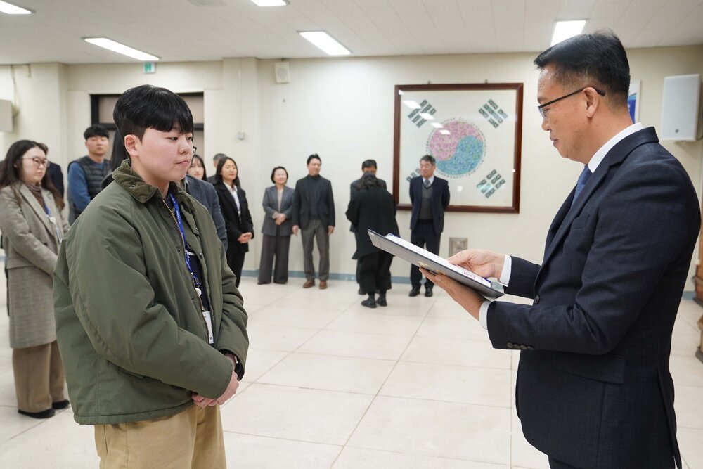 2026년 상반기 4급이하 정기인사로 정장을 입은 남성이 실내 행사장에서 한 남성에게 상장을 전달하며 내용을 읽고 있고, 남성은 카키색 짧은 패딩을 입고 두 손을 모은 상태로 서 있으며 뒤쪽으로 참석자들이 줄지어 서서 행사를 지켜보고 있다.