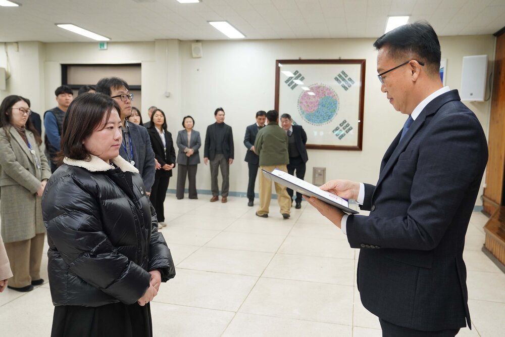2026년 상반기 4급이하 정기인사로 정장을 입은 남성이 실내 행사장에서 한 여성에게 상장을 전달하며 내용을 읽고 있고, 여성은 검은색 짧은 패딩을 입고 두 손을 모은 상태로 서 있으며 뒤쪽으로 참석자들이 줄지어 서서 행사를 지켜보고 있다.
