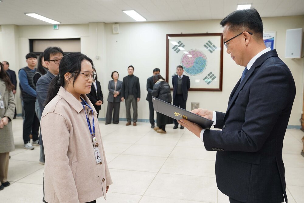 2026년 상반기 4급이하 정기인사로 정장을 입은 남성이 실내 행사장에서 한 여성에게 상장을 전달하며 내용을 읽고 있고, 여성은 밝은 베이지색 스웨이드 자켓을 입고 사원증을 착용한 채 두 손을 내린 상태로 서 있으며 뒤쪽에는 참석자들이 줄지어 서서 행사를 지켜보고 있다.