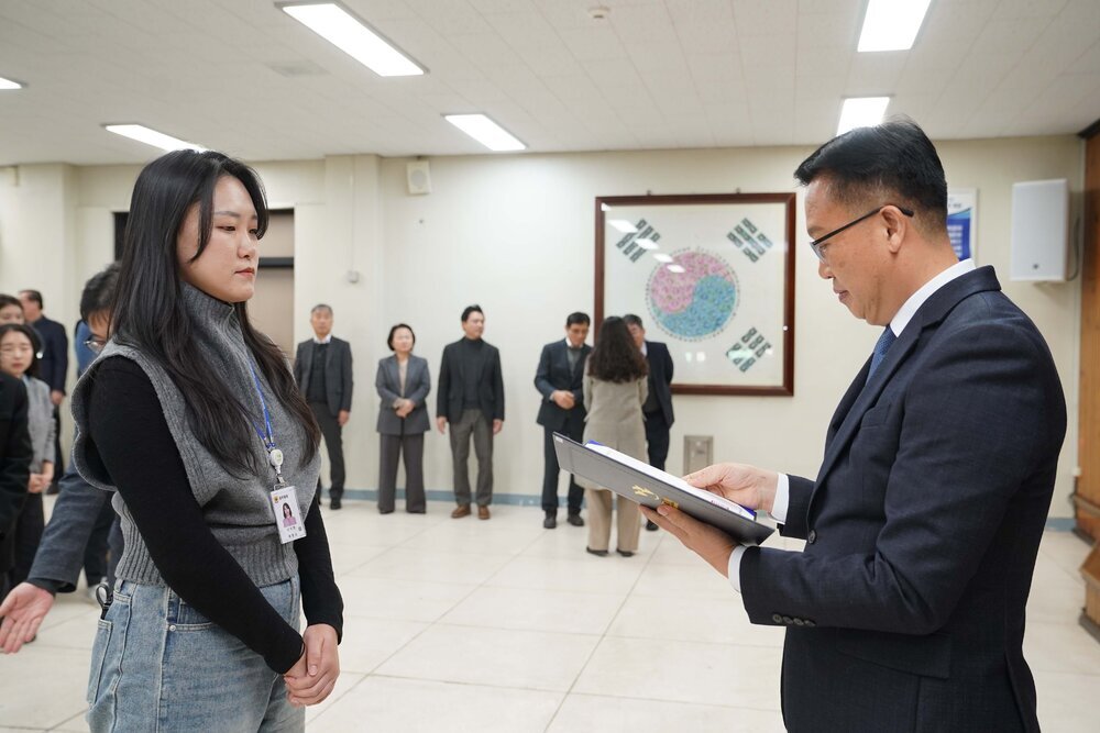2026년 상반기 4급이하 정기인사로 정장을 입은 남성이 실내 행사장에서 한 여성에게 상장을 전달하며 내용을 읽고 있고, 여성은 긴 생머리에 검은생 티에 회색 목까지올라오는 베스트를 입고 사원증을 착용한 채 두 손을 모은 상태로 서 있으며 뒤쪽에는 참석자들이 줄지어 서서 행사를 지켜보고 있다.