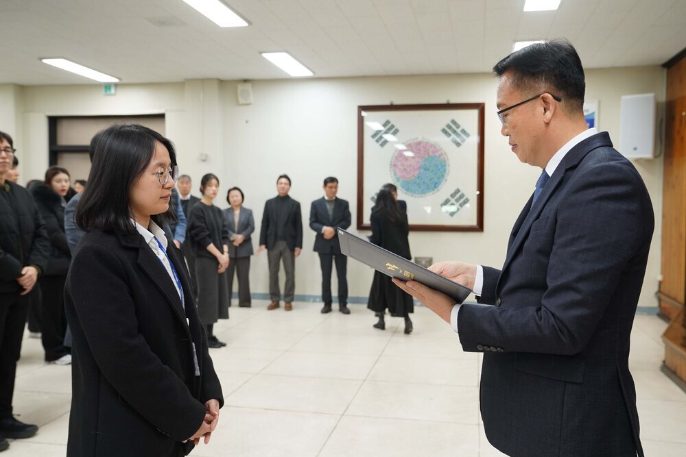 2026년 상반기 4급이하 정기인사로 정장을 입은 남성이 실내 행사장에서 한 여성에게 상장을 전달하며 내용을 읽고 있고, 여성은 어꺠 까지 오는 단발머리에 흰셔츠 검은색 자켓을 입고 사원증을 착용한 채 두 손을 모은 상태로 서 있으며 뒤쪽에는 참석자들이 줄지어 서서 행사를 지켜보고 있다.