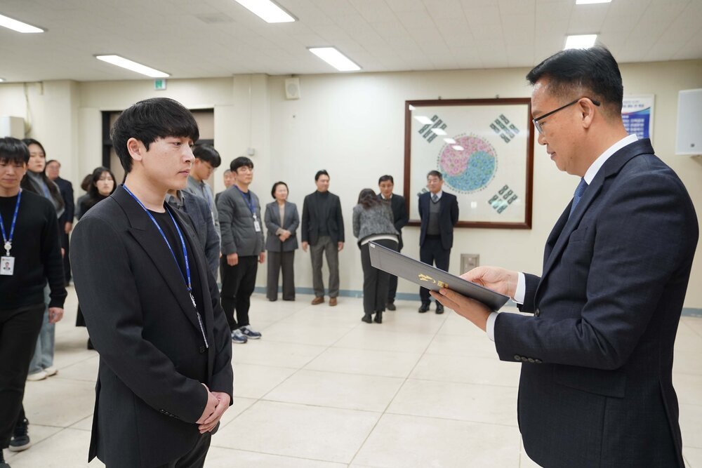 2026년 상반기 4급이하 정기인사로 정장을 입은 남성이 실내 행사장에서 한 남성에게 상장을 전달하며 내용을 읽고 있고, 남성은 검은 정장을 입은 채 두 손을 모은 상태로 서 있으며 뒤쪽에는 참석자들이 줄지어 서서 행사를 지켜보고 있다.