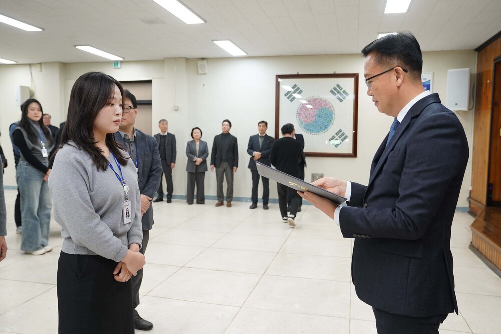 2026년 상반기 4급이하 정기인사로 정장을 입은 남성이 실내 행사장에서 한 여성에게 상장을 전달하며 내용을 읽고 있고, 여성은 회색 상의를 입고 두 손을 모은 상태로 서 있으며 뒤쪽에는 참석자들이 줄지어 서서 행사를 지켜보고 있다.