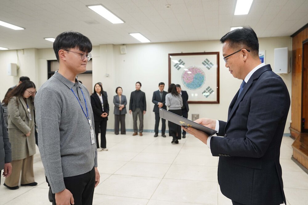 2026년 상반기 4급이하 정기인사로 정장을 입은 남성이 실내 행사장에서 한 남성에게 상장을 전달하며 내용을 읽고 있고, 남성은 회색 니트를 입은 채 두 손을 모은 상태로 서 있으며 뒤쪽에는 참석자들이 줄지어 서서 행사를 지켜보고 있다.