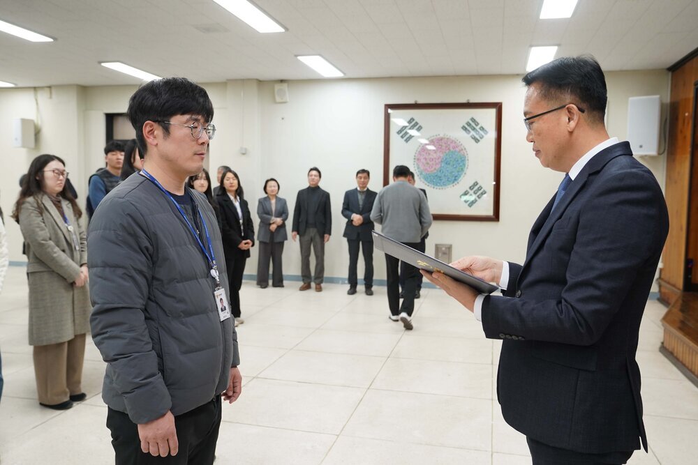 2026년 상반기 4급이하 정기인사로 정장을 입은 남성이 실내 행사장에서 한 남성에게 상장을 전달하며 내용을 읽고 있고, 남성은 회색 패딩을 입은 채 두 손을 모은 상태로 서 있으며 뒤쪽에는 참석자들이 줄지어 서서 행사를 지켜보고 있다.
