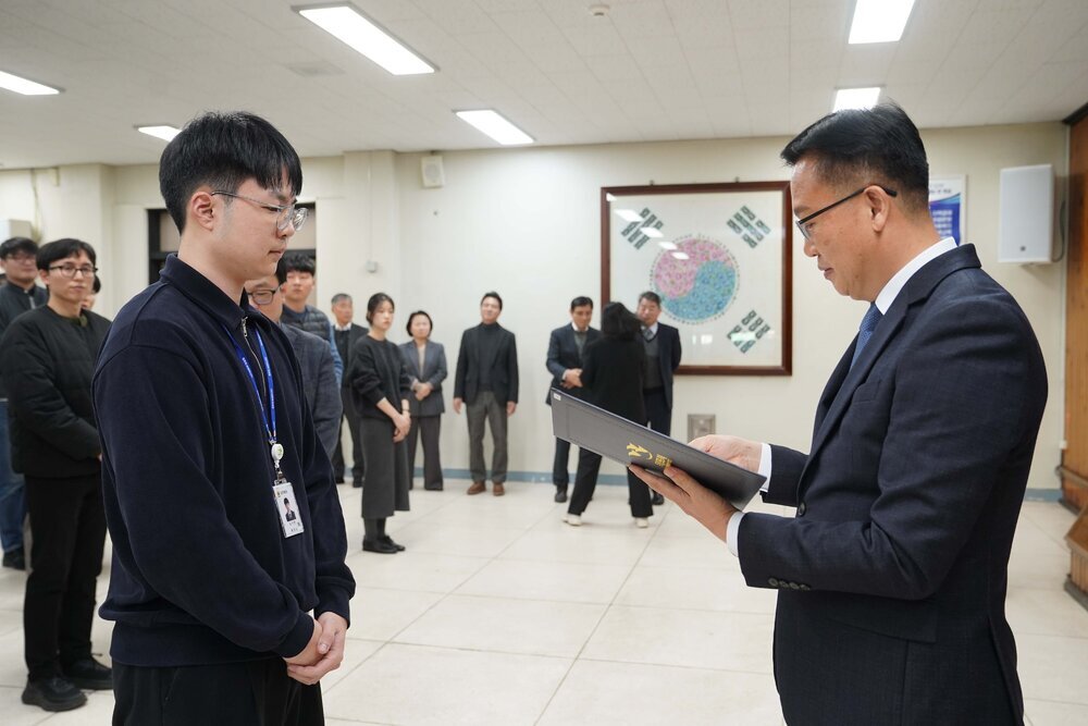 2026년 상반기 4급이하 정기인사로 정장을 입은 남성이 실내 행사장에서 한 남성에게 상장을 전달하며 내용을 읽고 있고, 남성은 검은 상의를 입고 사원증을 착용한 채 두 손을 모은 상태로 서 있으며 뒤쪽에는 참석자들이 줄지어 서서 행사를 지켜보고 있다.