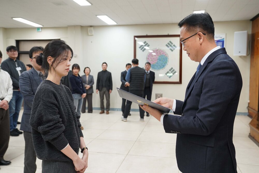 2026년 상반기 4급이하 정기인사로 정장을 입은 남성이 실내 행사장에서 한 여성에게 상장을 전달하며 내용을 읽고 있고, 여성은 검은 니트를 입고 두 손을 모은 상태로 서 있으며 뒤쪽에는 참석자들이 줄지어 서서 행사를 지켜보고 있다.