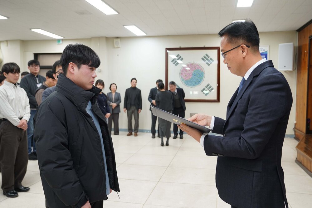 2026년 상반기 4급이하 정기인사로 정장을 입은 남성이 실내 행사장에서 한 남성에게 상장을 전달하며 내용을 읽고 있고, 남성은 검은 점퍼를 입은 채 두 손을 모은 상태로 서 있으며 뒤쪽에는 참석자들이 줄지어 서서 행사를 지켜보고 있다.