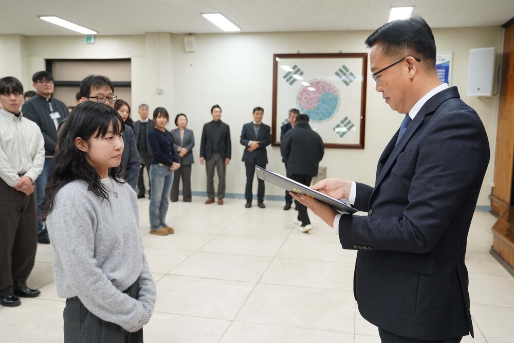 2026년 상반기 4급이하 정기인사로 정장을 입은 남성이 실내 행사장에서 한 여성에게 상장을 전달하며 내용을 읽고 있고, 여성은 회색 니트를 입고 두 손을 모은 상태로 서 있으며 뒤쪽에는 참석자들이 줄지어 서서 행사를 지켜보고 있다.