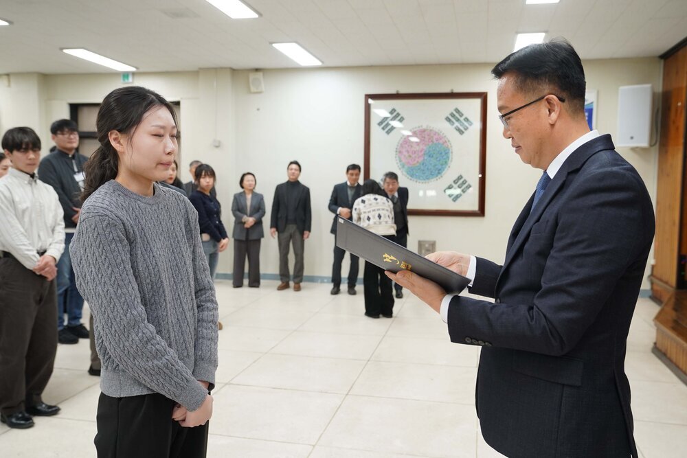 2026년 상반기 4급이하 정기인사로 정장을 입은 남성이 실내 행사장에서 한 여성에게 상장을 전달하며 내용을 읽고 있고, 여성은 회색 니트를 입고 두 손을 모은 상태로 서 있으며 뒤쪽에는 참석자들이 줄지어 서서 행사를 지켜보고 있다.