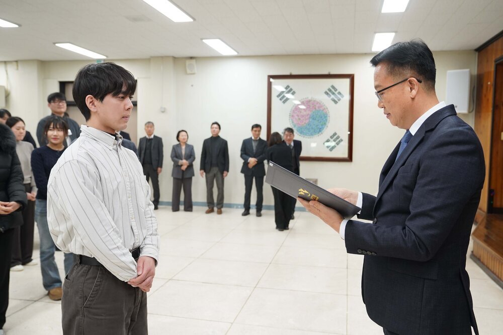 2026년 상반기 4급이하 정기인사로 정장을 입은 남성이 실내 행사장에서 한 남성에게 상장을 전달하며 내용을 읽고 있고, 남성은 줄무늬 셔츠를 입은 채 두 손을 모은 상태로 서 있으며 뒤쪽에는 참석자들이 줄지어 서서 행사를 지켜보고 있다.