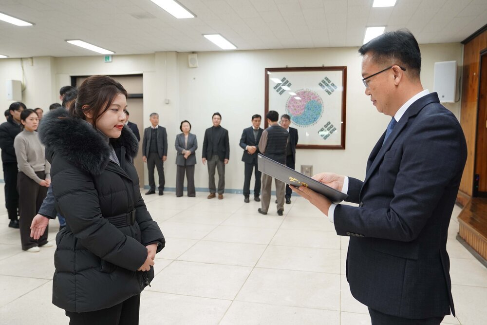 2026년 상반기 4급이하 정기인사로 정장을 입은 남성이 실내 행사장에서 한 여성에게 상장을 전달하며 내용을 읽고 있고, 여성은 검은 패딩 코트를 입고 두 손을 모은 상태로 서 있으며 뒤쪽에는 참석자들이 줄지어 서서 행사를 지켜보고 있다.