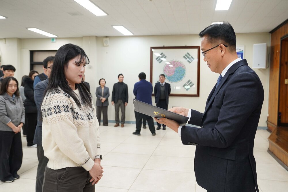 2026년 상반기 4급이하 정기인사로 정장을 입은 남성이 실내 행사장에서 한 여성에게 상장을 전달하며 내용을 읽고 있고, 여성은 무늬가 있는 니트를 입고 두 손을 모은 상태로 서 있으며 뒤쪽에는 참석자들이 줄지어 서서 행사를 지켜보고 있다.
