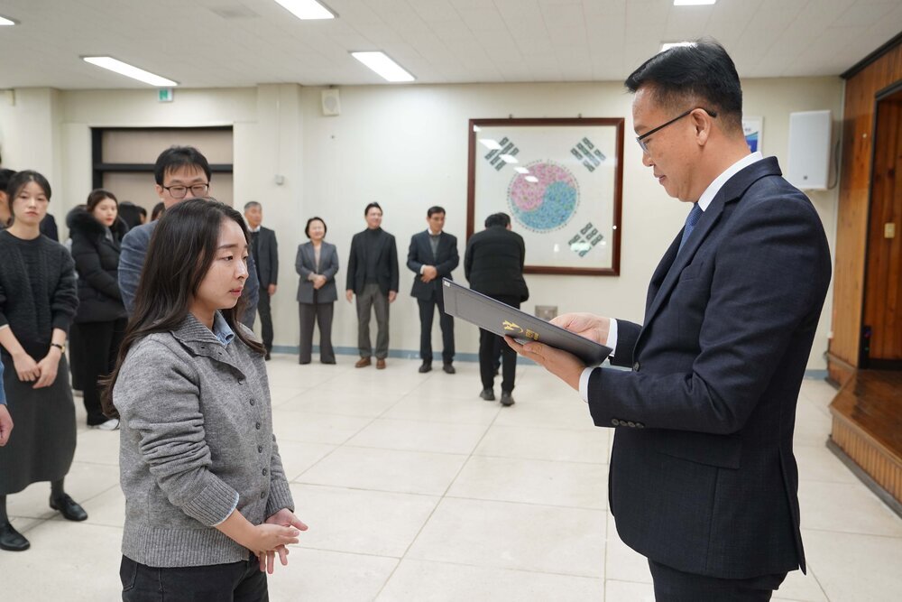 2026년 상반기 4급이하 정기인사로 정장을 입은 남성이 실내 행사장에서 한 여성에게 상장을 전달하며 내용을 읽고 있고, 여성은 회색 니트를 입고 두 손을 모은 상태로 서 있으며 뒤쪽에는 참석자들이 줄지어 서서 행사를 지켜보고 있다.