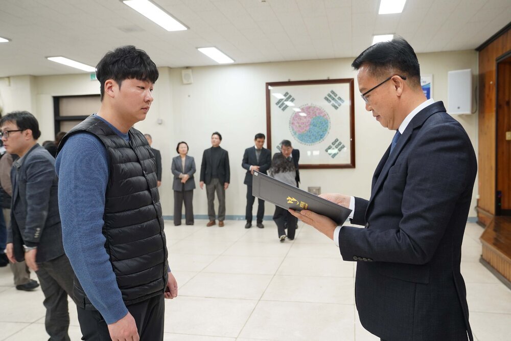 2026년 상반기 4급이하 정기인사로 정장을 입은 남성이 실내 행사장에서 한 남성에게 상장을 전달하며 내용을 읽고 있고, 남성은 검은 패딩 조끼를 입은 채 두 손을 모은 상태로 서 있으며 뒤쪽에는 참석자들이 줄지어 서서 행사를 지켜보고 있다.